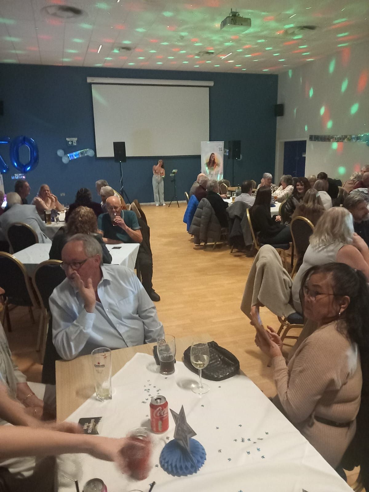 Party in a hall: People seated at tables watching a performer on stage under disco lights.
