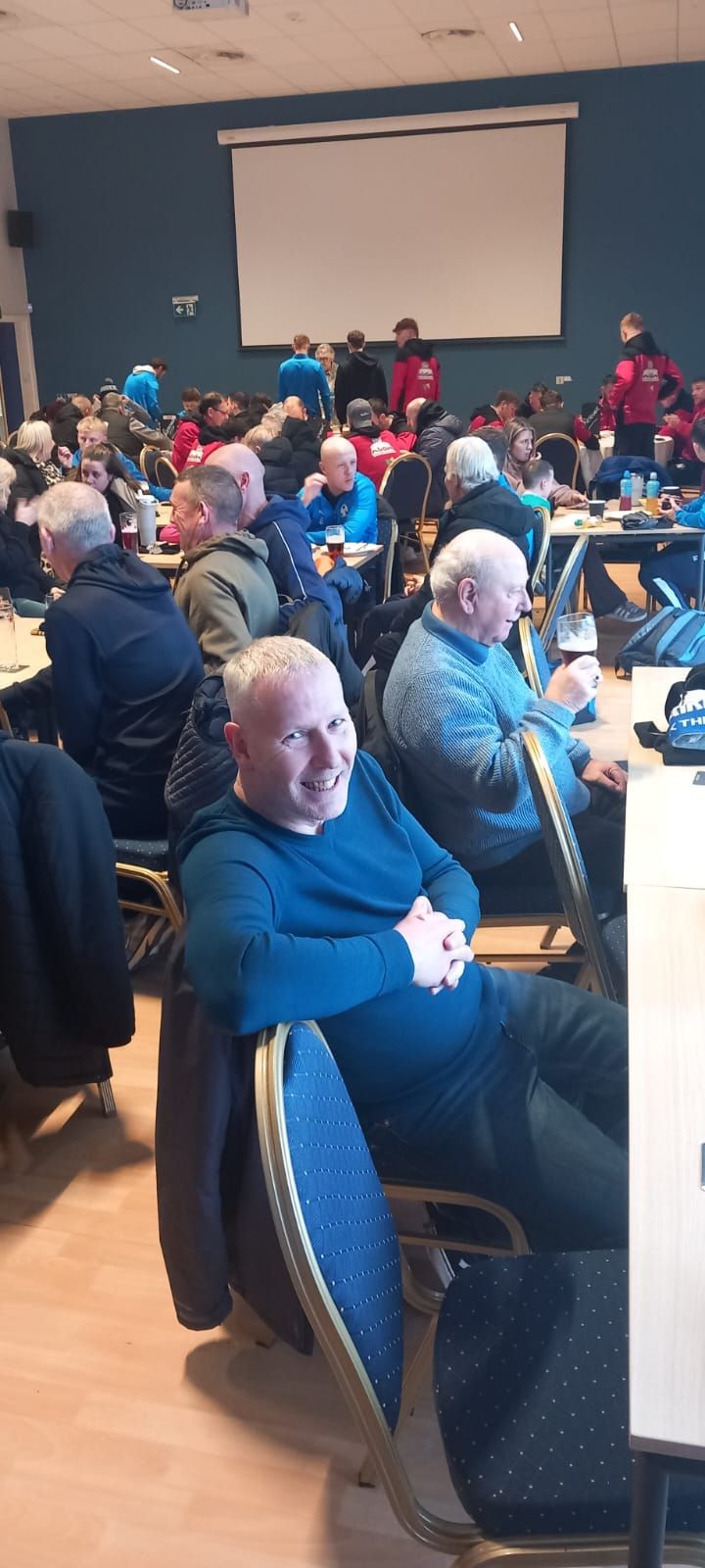 Man with crossed arms smiles in a room with a crowd. Others are seated at tables near a projector screen.