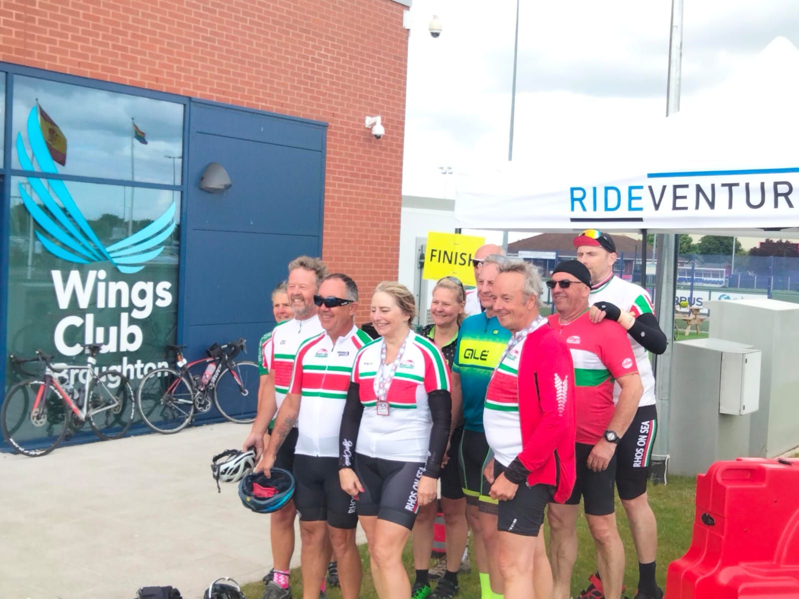 Group of cyclists pose in front of a building with a “Wings Club” sign after a Rideventure Brighton event.