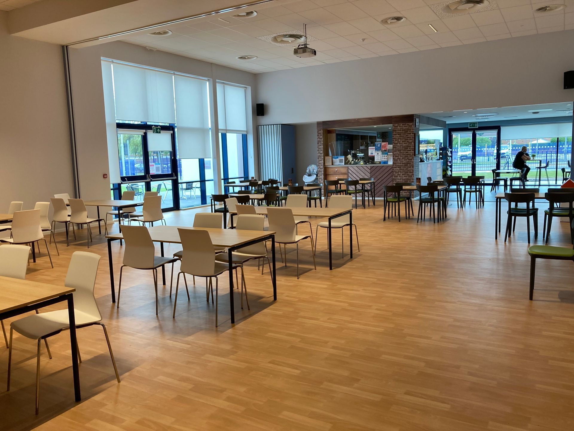 A large, open room with tables and chairs, a bar, and windows overlooking a sports field.