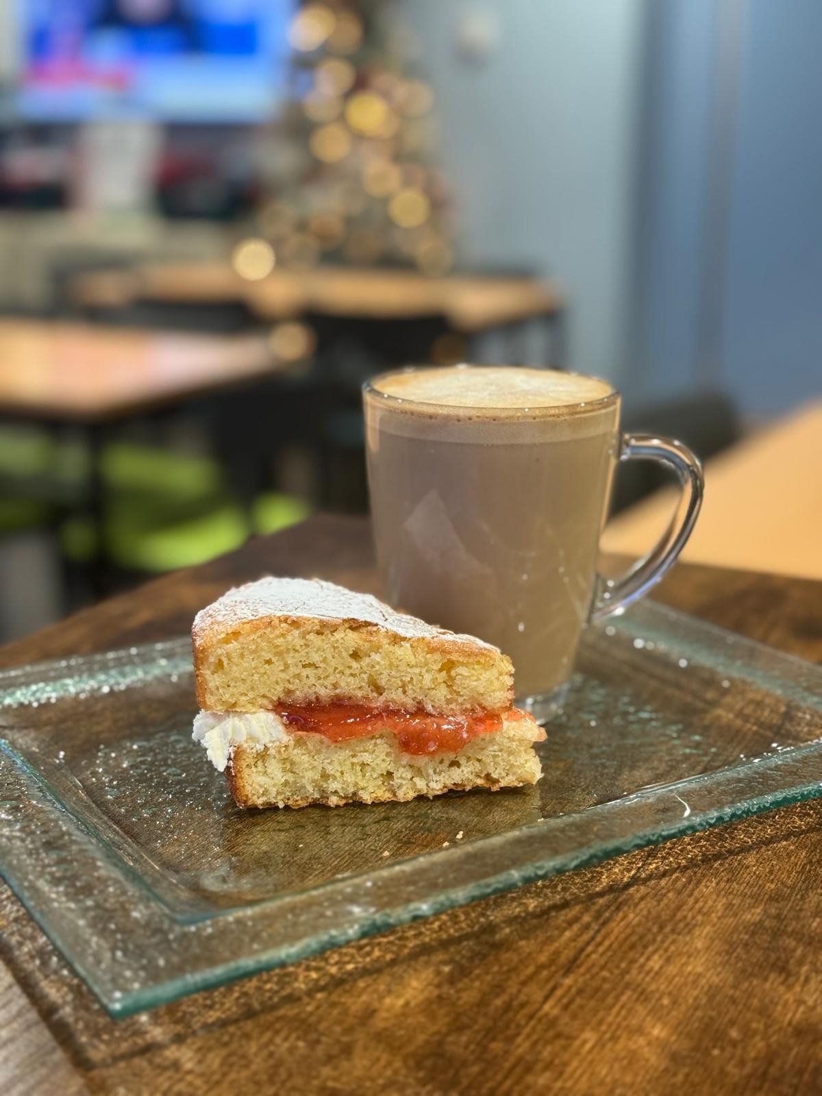 Slice of cake with jam and powdered sugar, next to a mug of coffee on a glass plate, on a wooden table.
