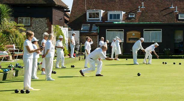 People in white attire playing lawn bowls on a green lawn, in front of a clubhouse.