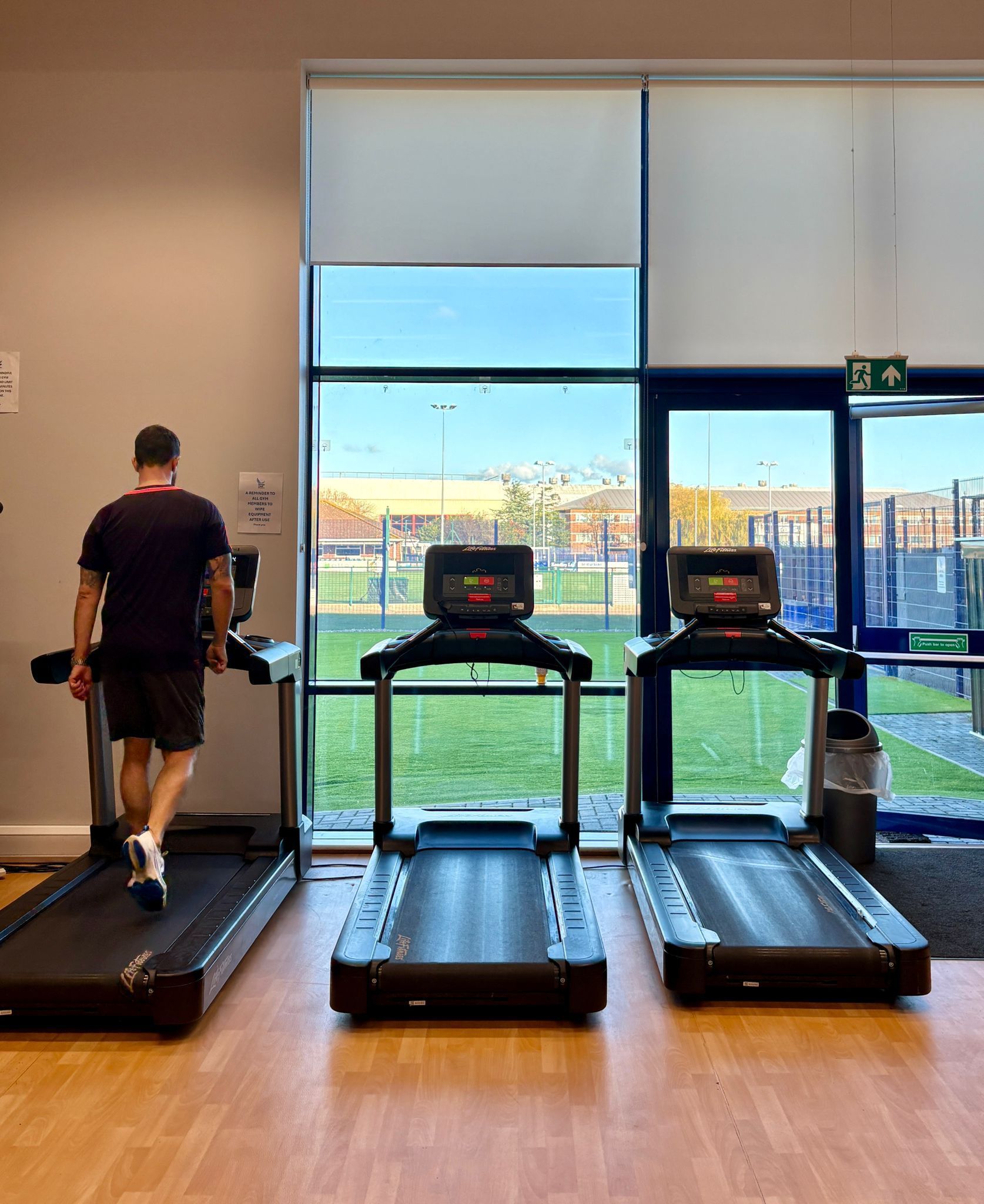 A person walks on a treadmill in a gym, with two empty treadmills beside a large window overlooking a field.