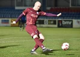 Soccer player in maroon uniform kicking a ball on a green field at a stadium.
