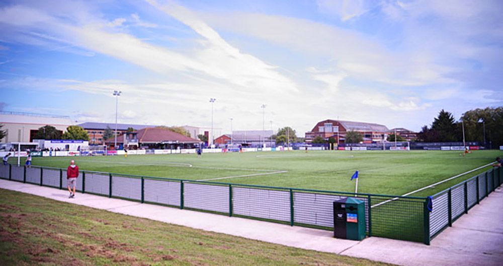 Green sports field enclosed by a low fence; buildings in the background under a blue sky.
