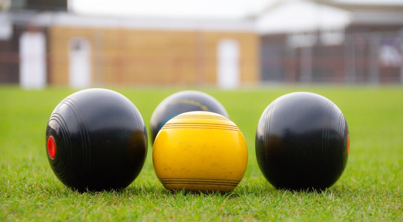 Lawn bowls: Four bowls (3 black, 1 yellow) on green grass, a wooden building in the background.