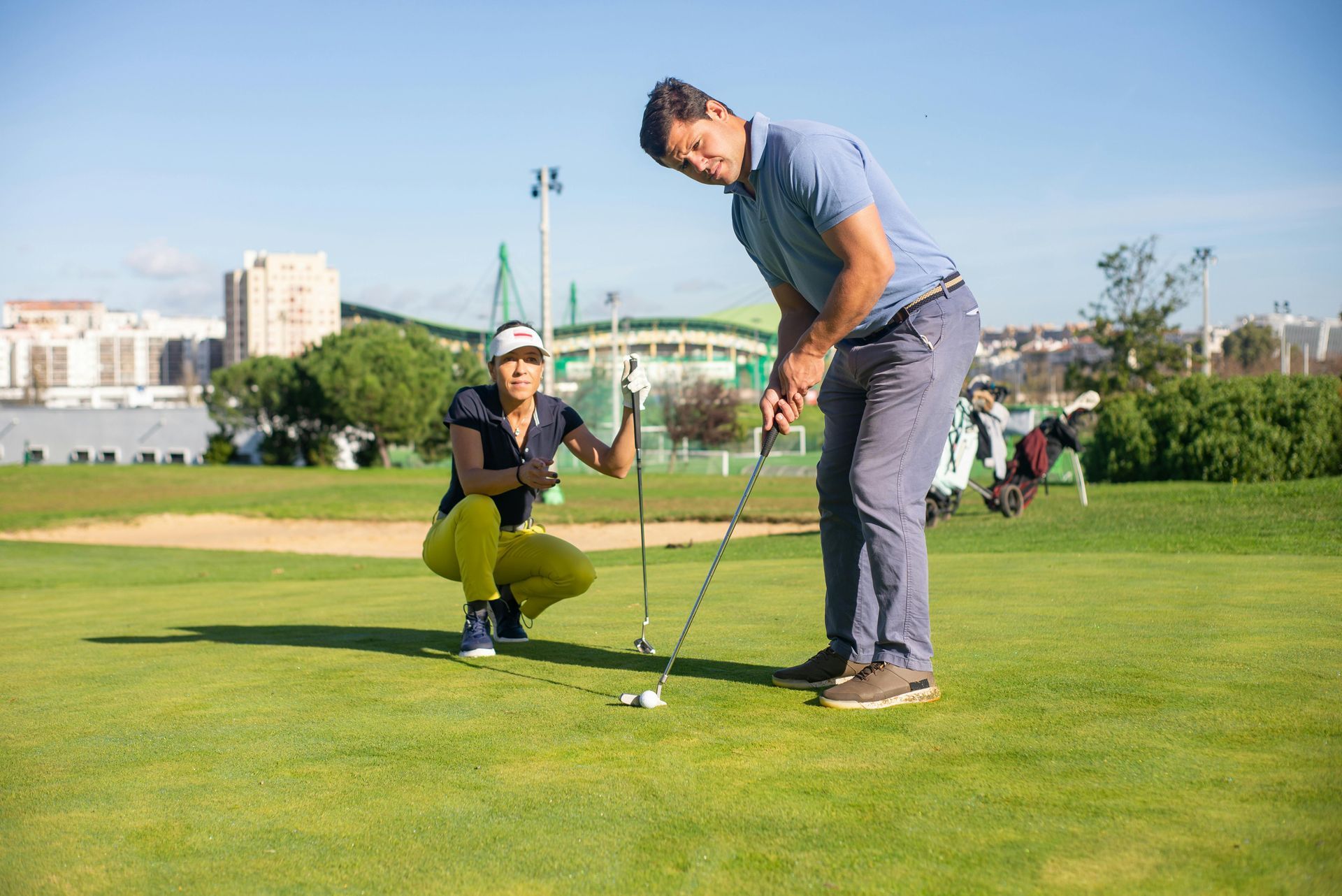 Man golfing, being watched by a woman kneeling on the grass. Green course, sunny day, city in the background.