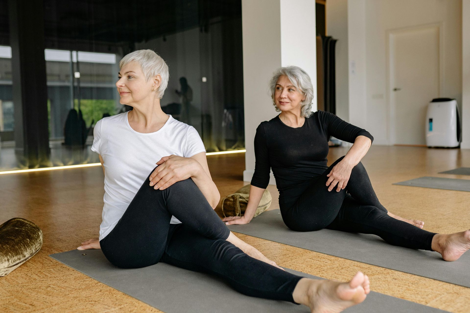 Two women doing seated spinal twist yoga pose on mats in a studio.