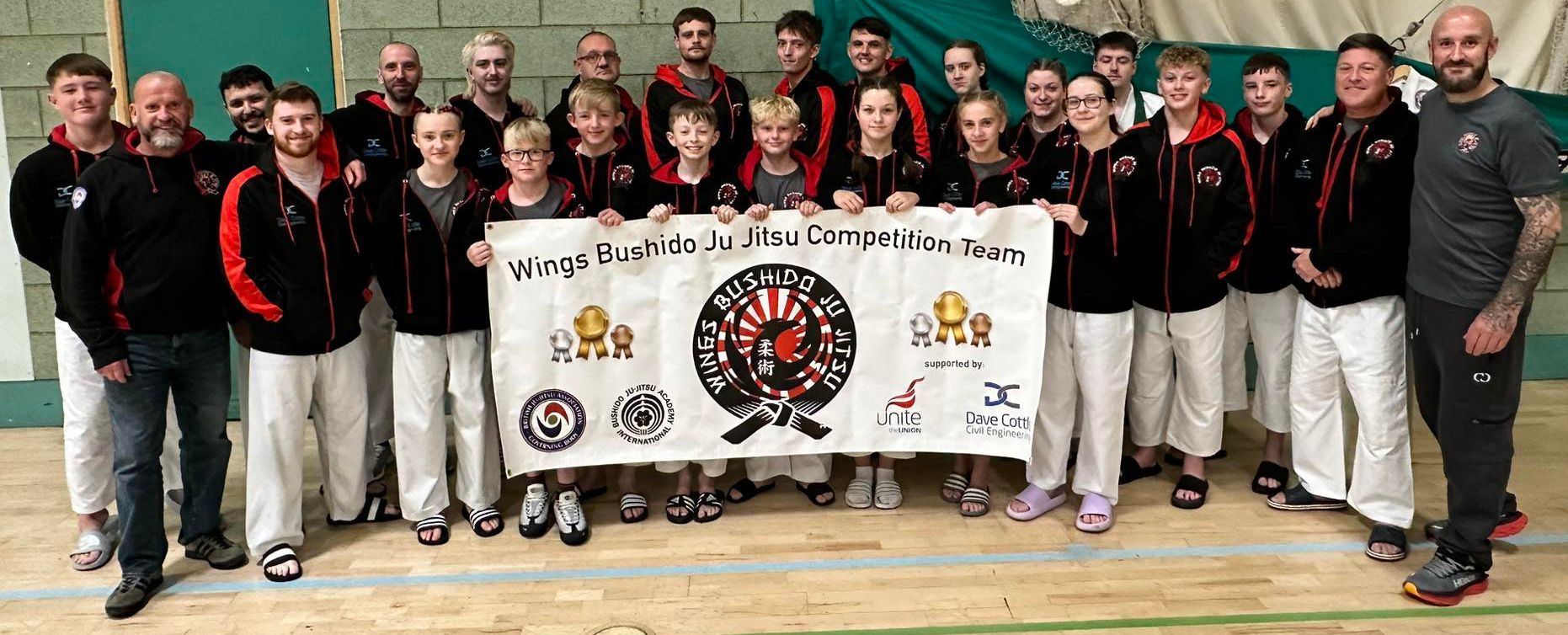Group of people holding a banner with a logo in a gym. Many wear black jackets with red trim and white pants.