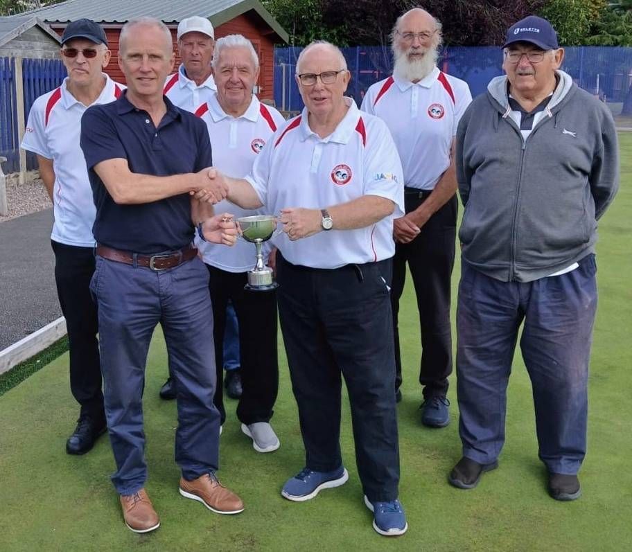 Group of men on a lawn, one receives a trophy. They wear white shirts, standing near a green bowling field.