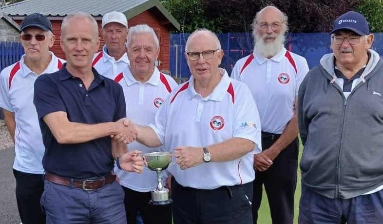 Group of men on a lawn; one shaking hands, another holding a trophy, others in matching white shirts.