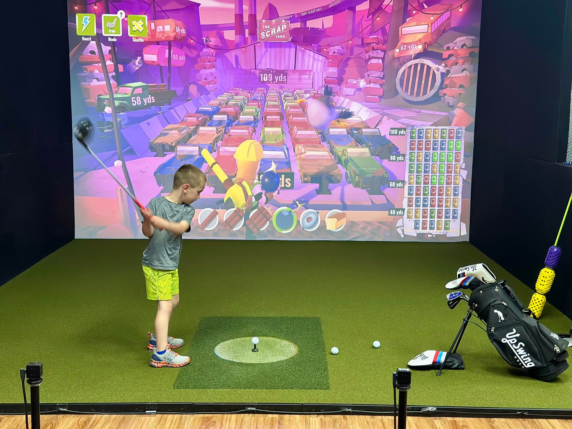 Child golfing in an indoor simulator, swinging at a virtual course with a large projected screen.