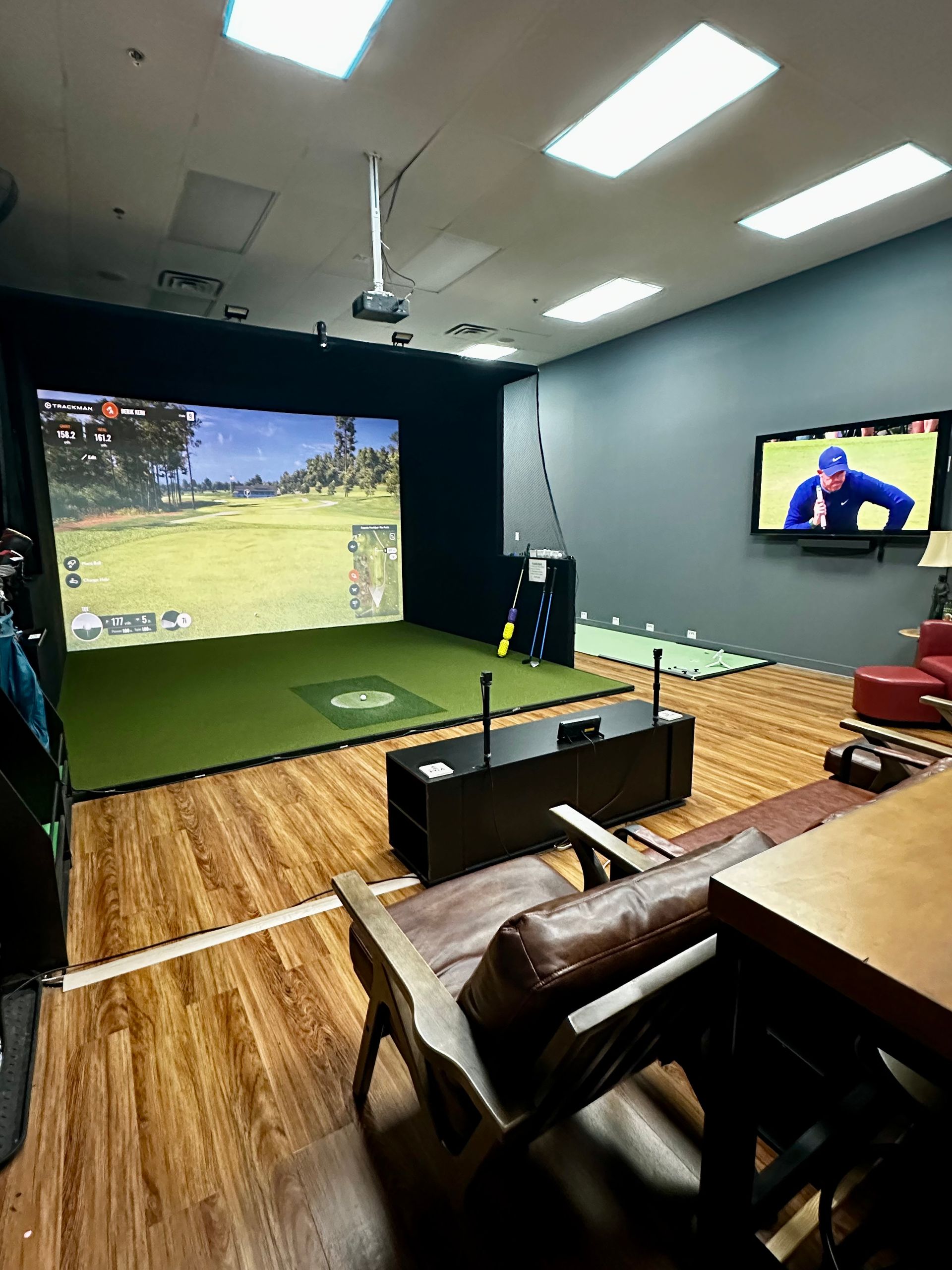 Indoor golf simulator room with large projection screen, lounge chairs, and wall-mounted TV showing a golfer