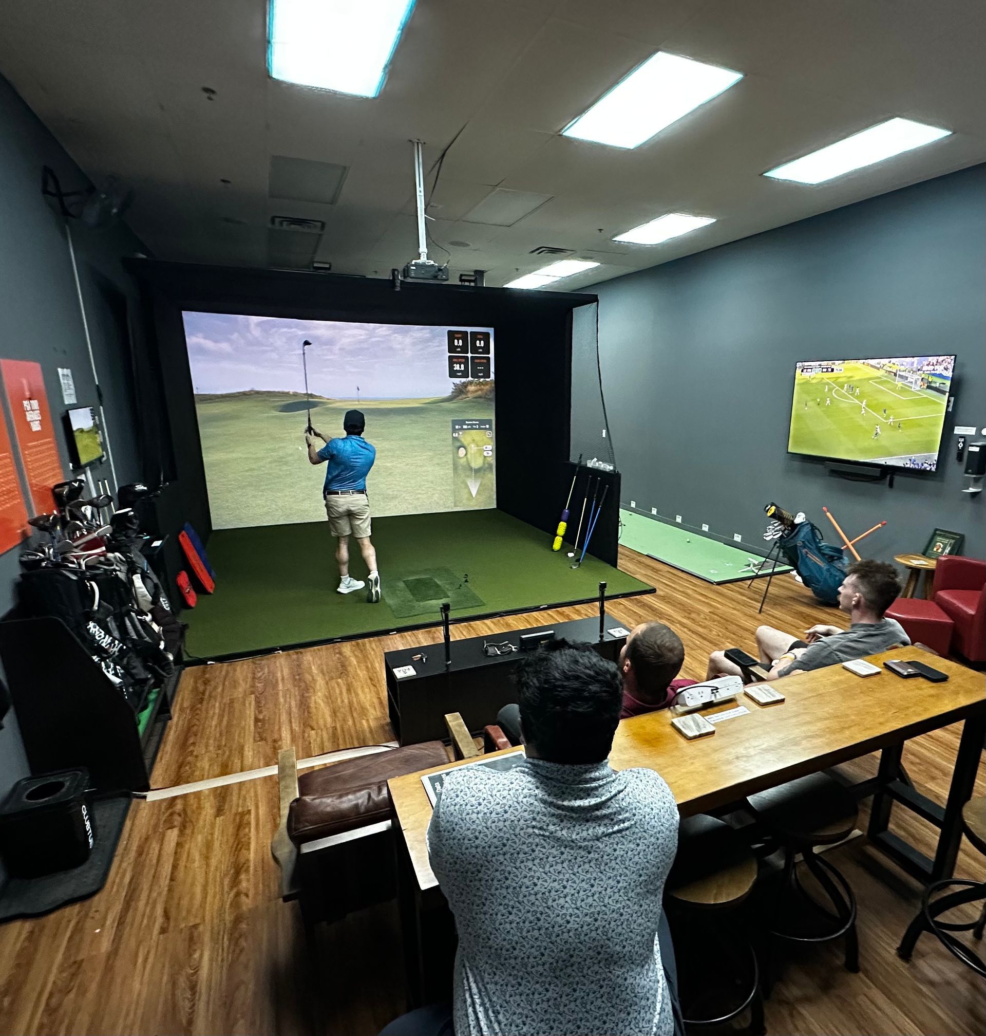 Indoor golf simulator room with two people watching a golfer swing at a projected course screen