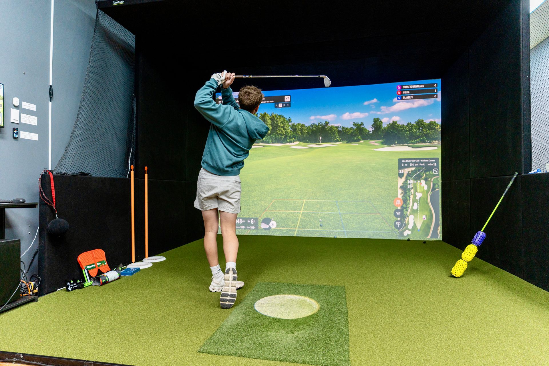 Person swinging a golf club in an indoor simulator bay with green turf and a large projection screen
