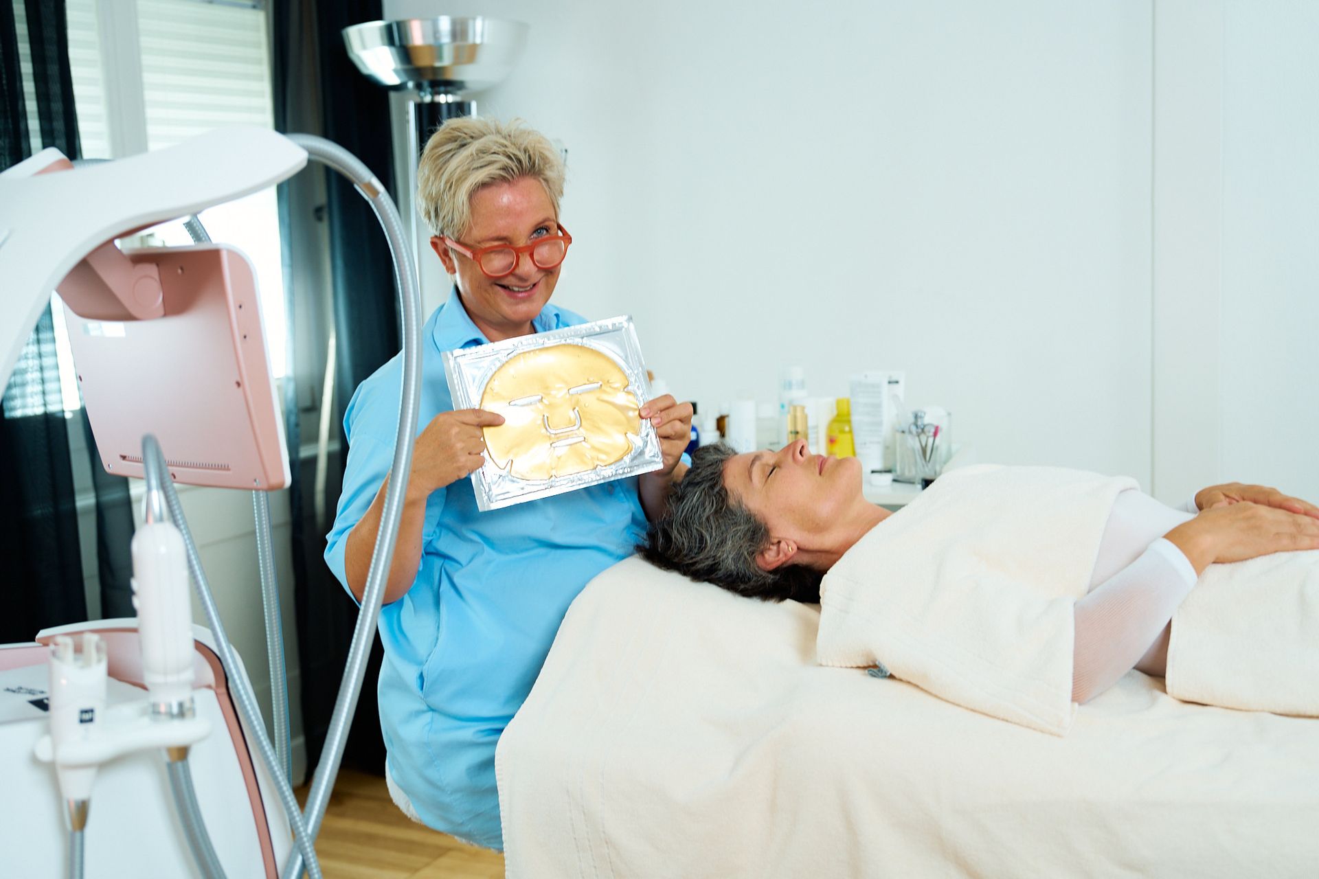 Eine Frau liegt auf einem Bett, während eine Kosmetikerin eine Goldene Gesichtsmaske in den Händen hält