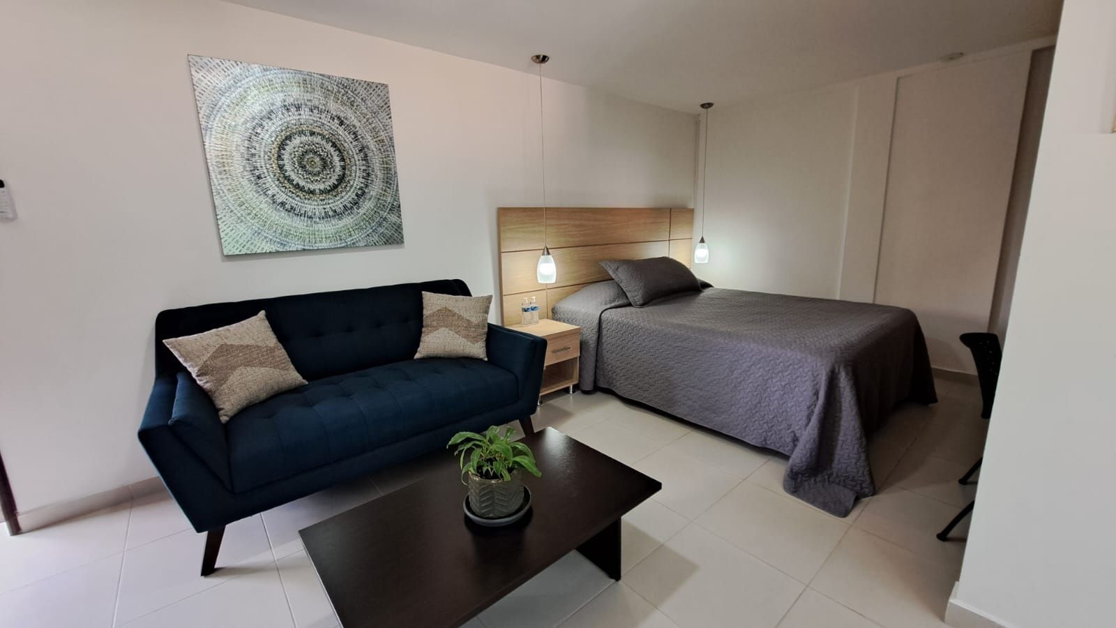 Habitación pequeña y moderna con sofá azul oscuro, mesa de centro, cama y cuadros en la pared.