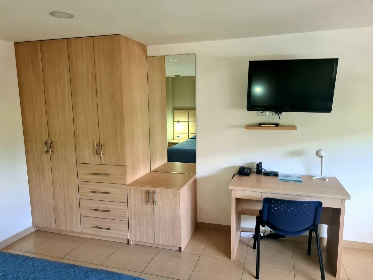 Habitación pequeña con armario de madera, escritorio, silla, televisor montado en la pared y suelo de baldosas.
