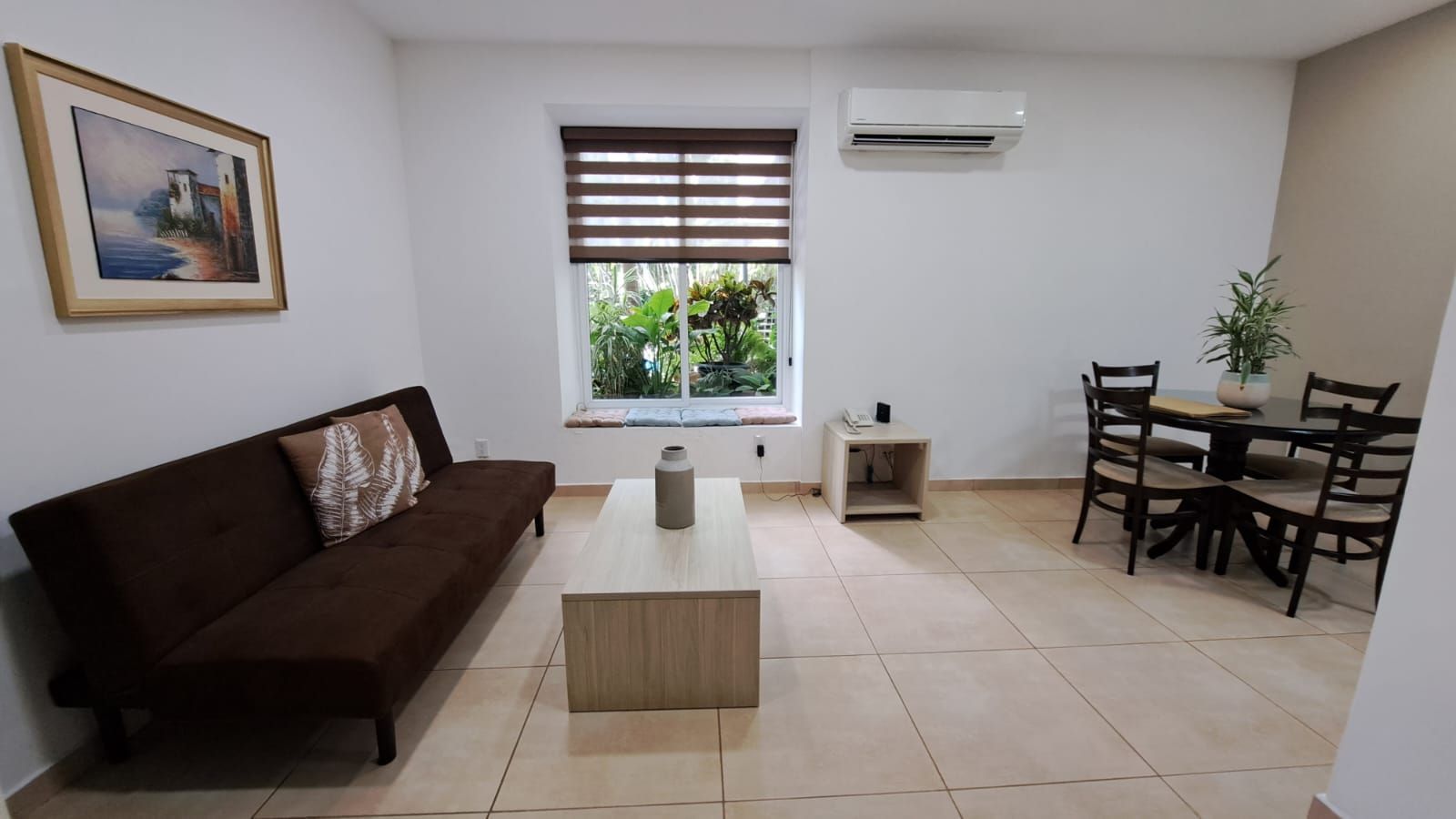 Sala de estar luminosa con sofá marrón, mesa de centro, juego de comedor, ventana y aire acondicionado mural.