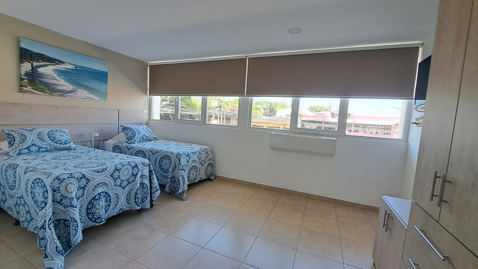 Habitación luminosa con dos camas estampadas en azul, suelo de baldosas y grandes ventanas con persianas marrones.