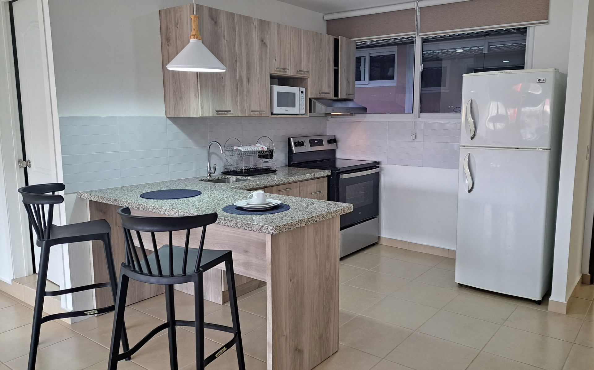 Cocina moderna con isla de granito, dos taburetes negros, estufa, microondas y refrigerador blanco.