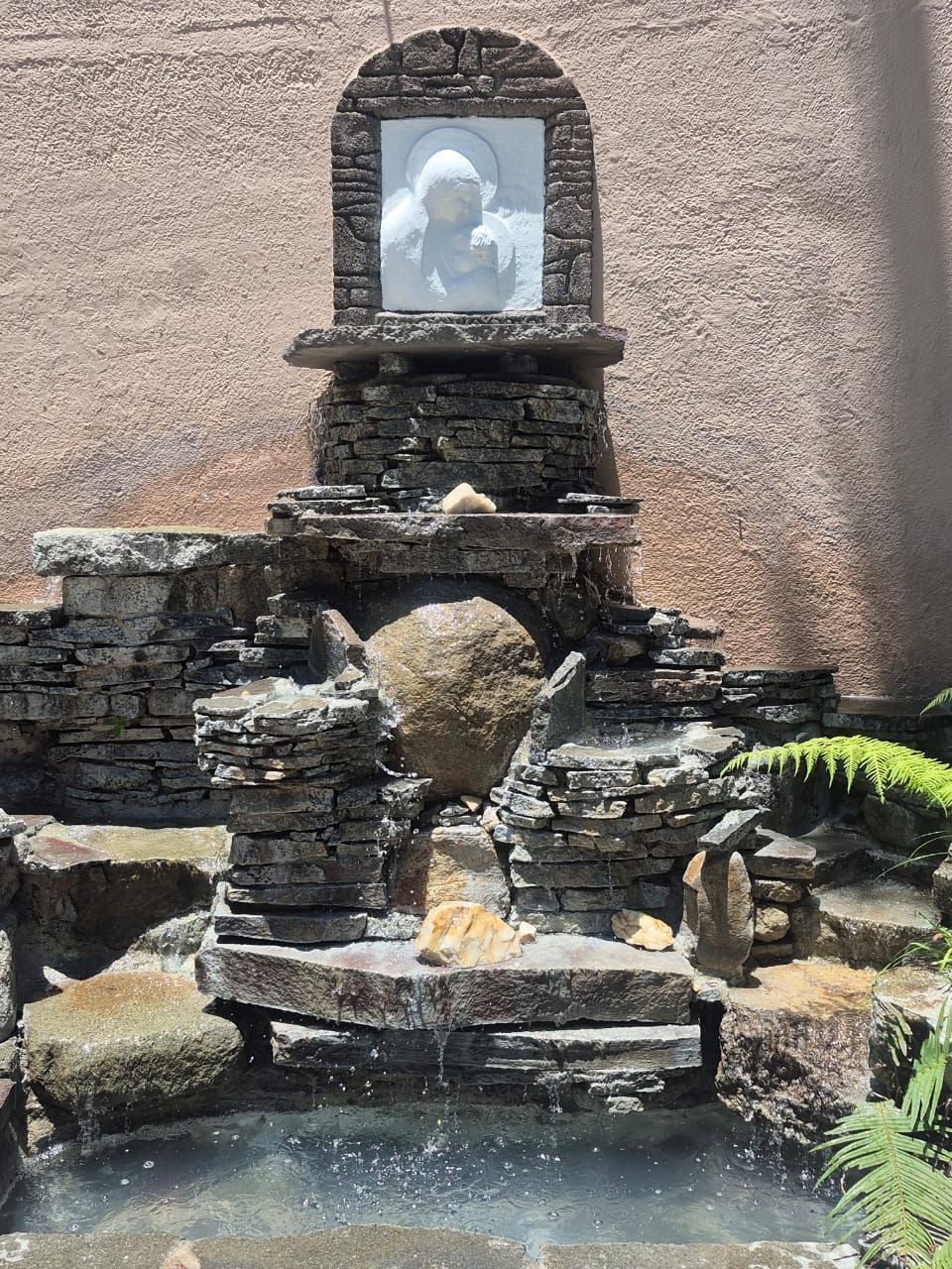 Fuente de jardín de piedra con una pequeña estatua en un nicho, agua que fluye hacia un estanque.