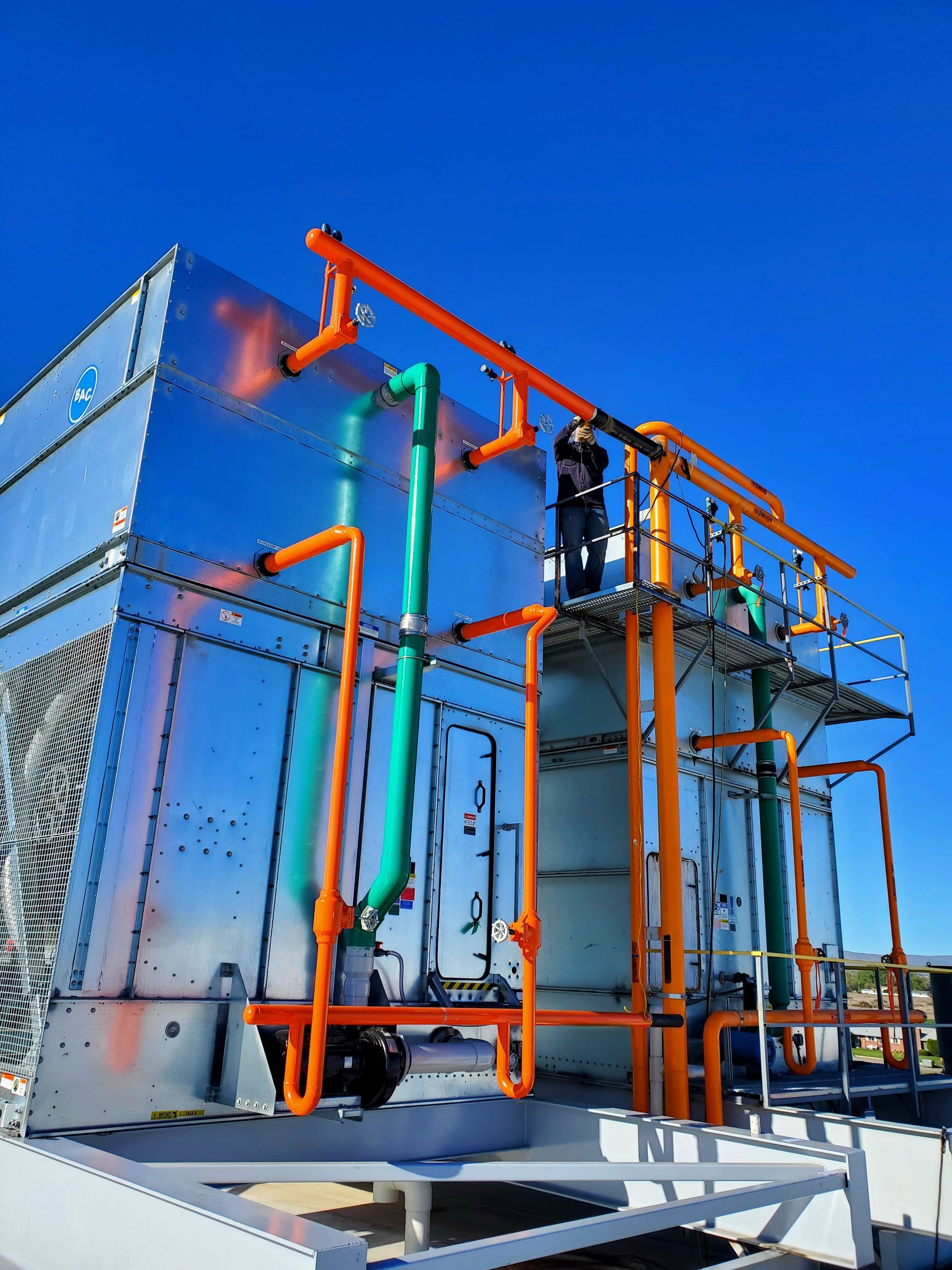 Cooling tower with orange pipes, green pipes, and silver panels against a blue sky.