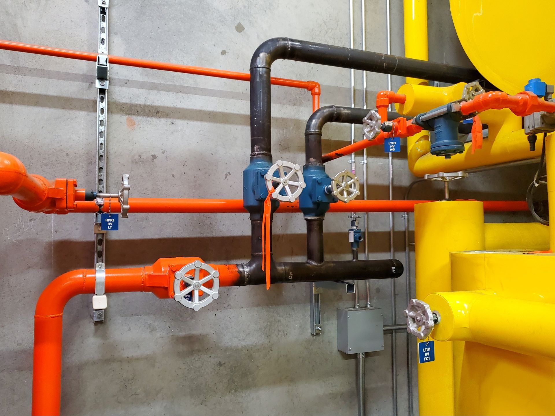 Industrial pipes and valves mounted on a wall, with bright orange, blue, and yellow piping.