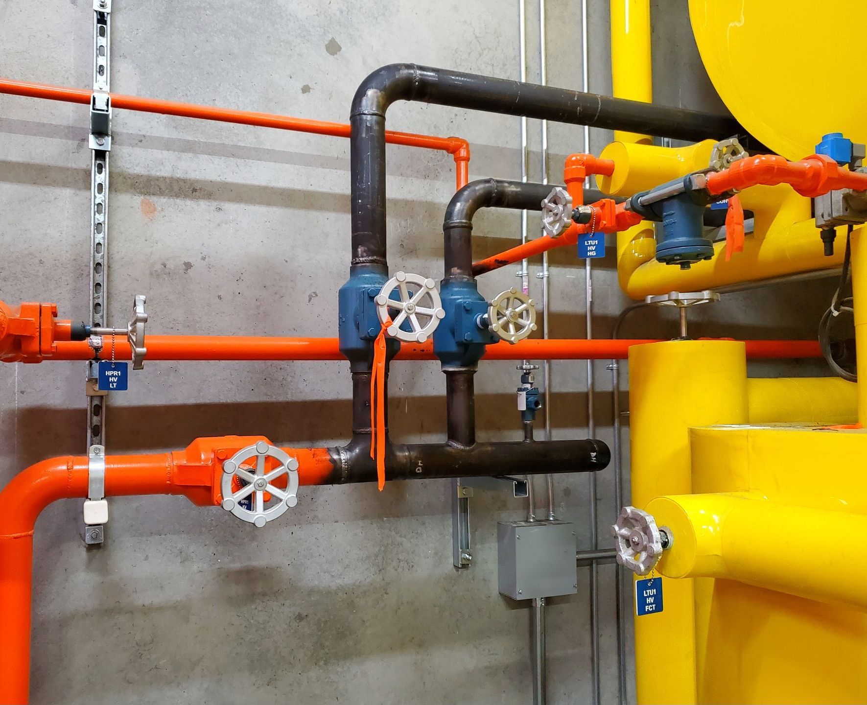 Orange and black pipes with blue valves against a concrete wall, near yellow machinery.