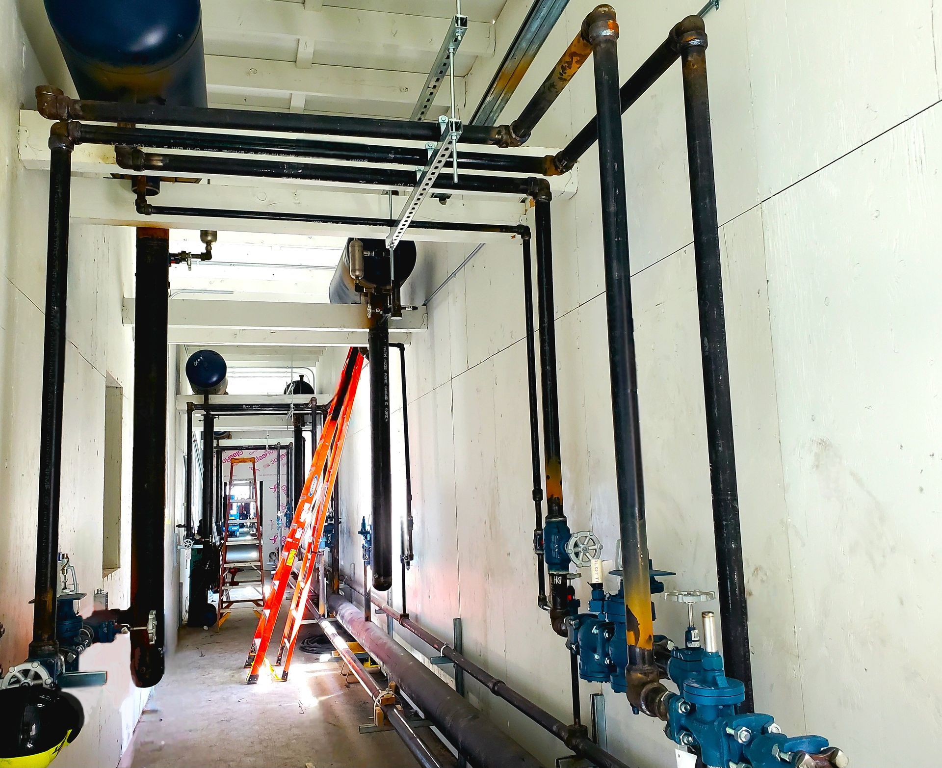 Narrow industrial corridor with exposed pipes, blue valves, and an orange ladder under construction to the left.