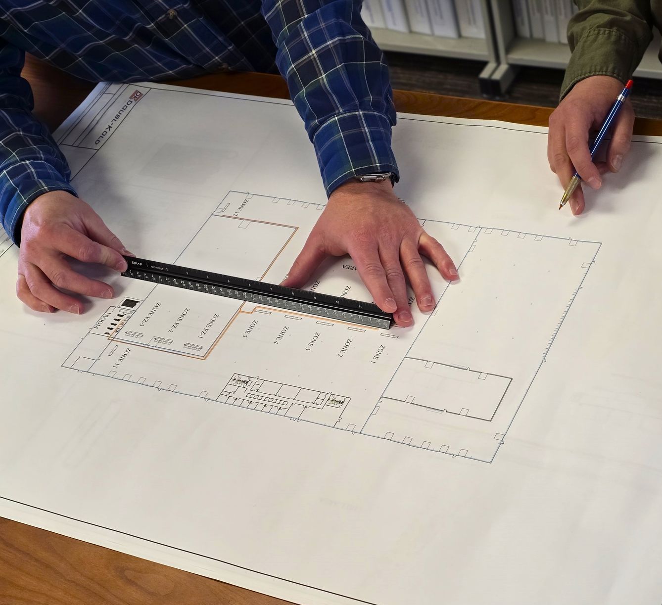 Two people's hands using a ruler and pencil on architectural blueprints.