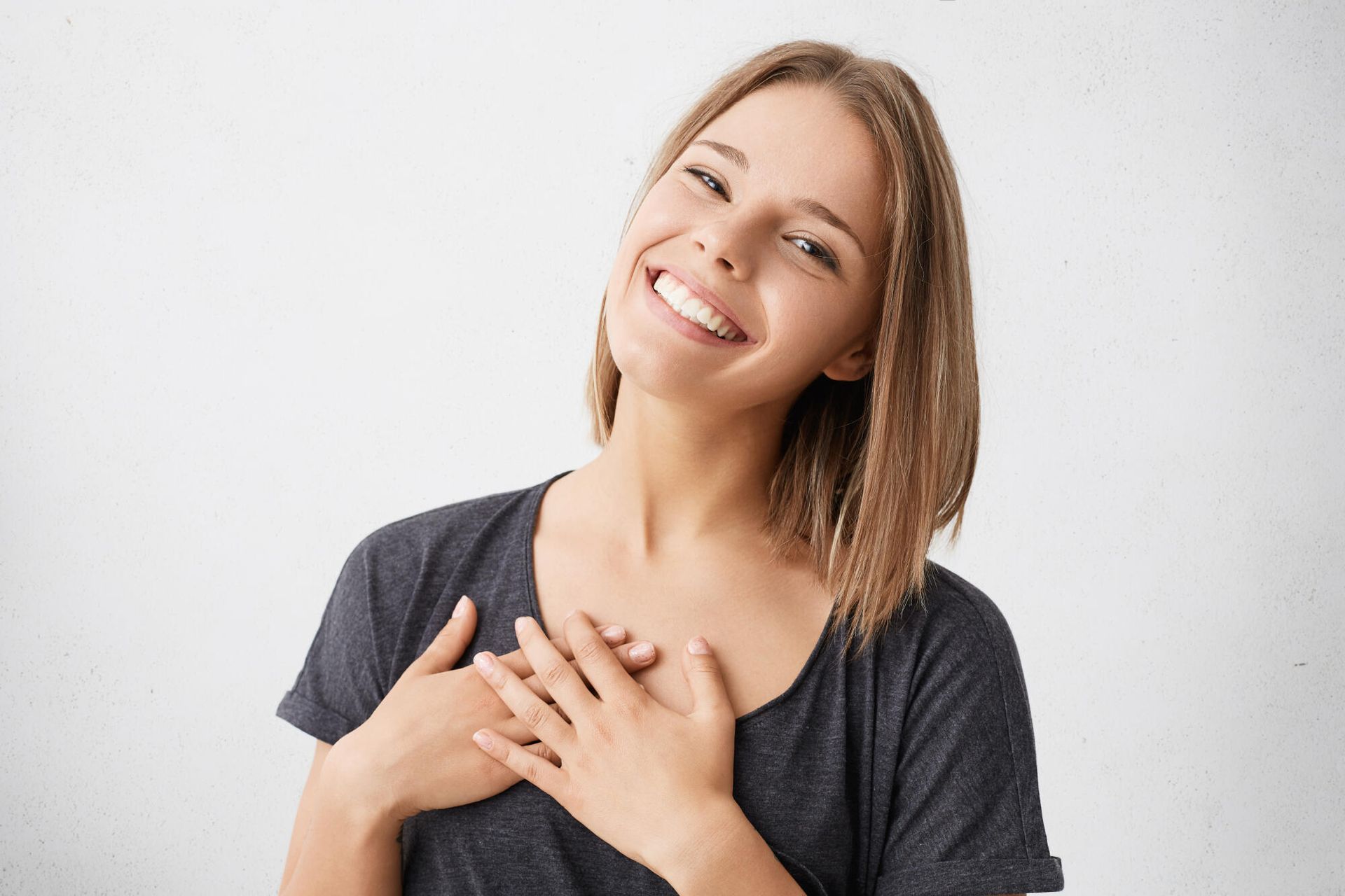 Smiling person with hands on chest, wearing a dark gray shirt against a light background