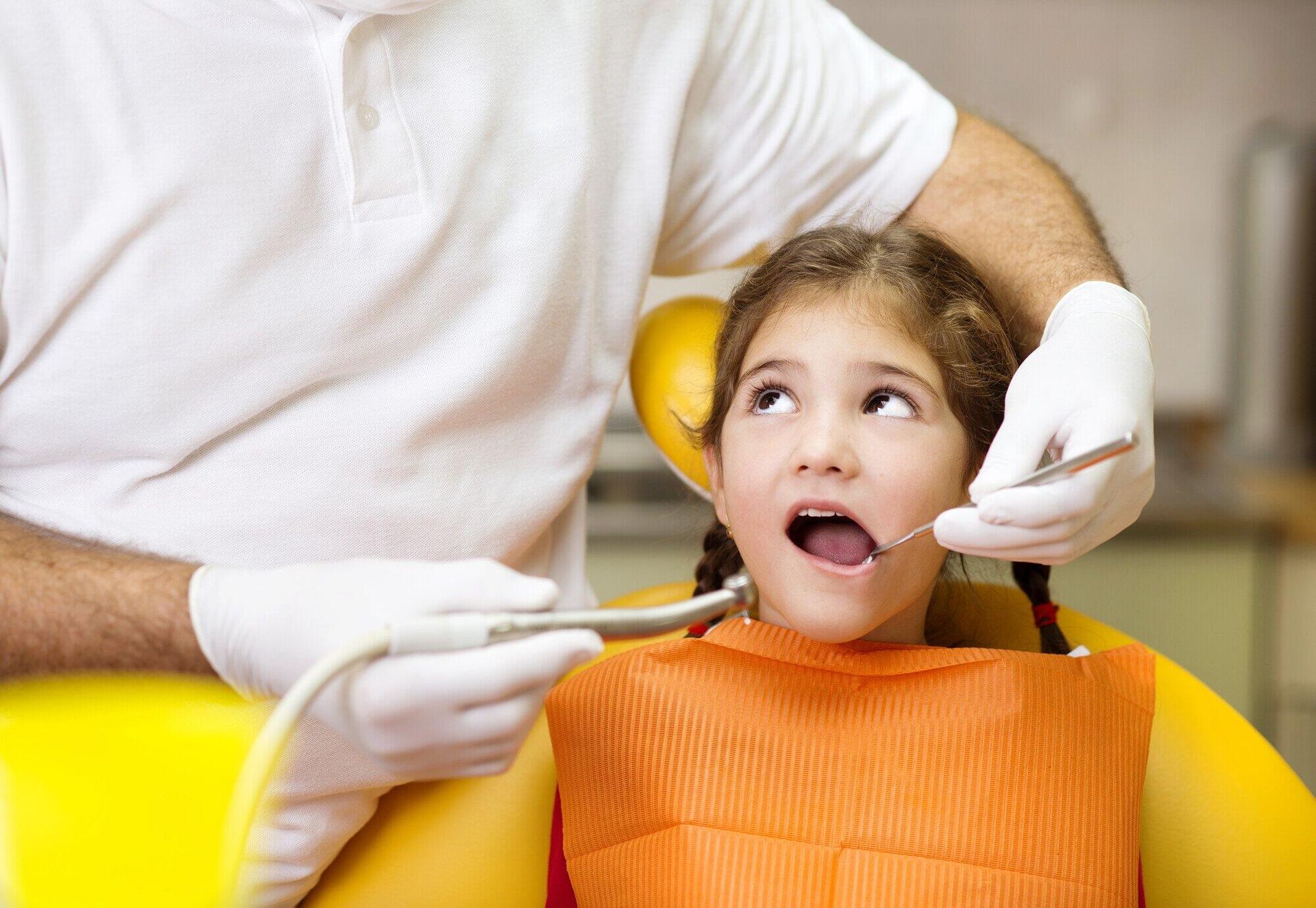 Dentist examining a young child in a yellow chair, child looking surprised, wearing an orange bib