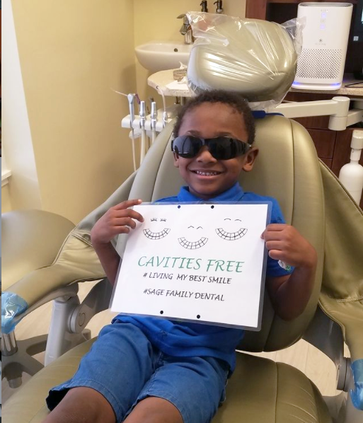 A young boy in a dental chair holding a sign that says cavities free