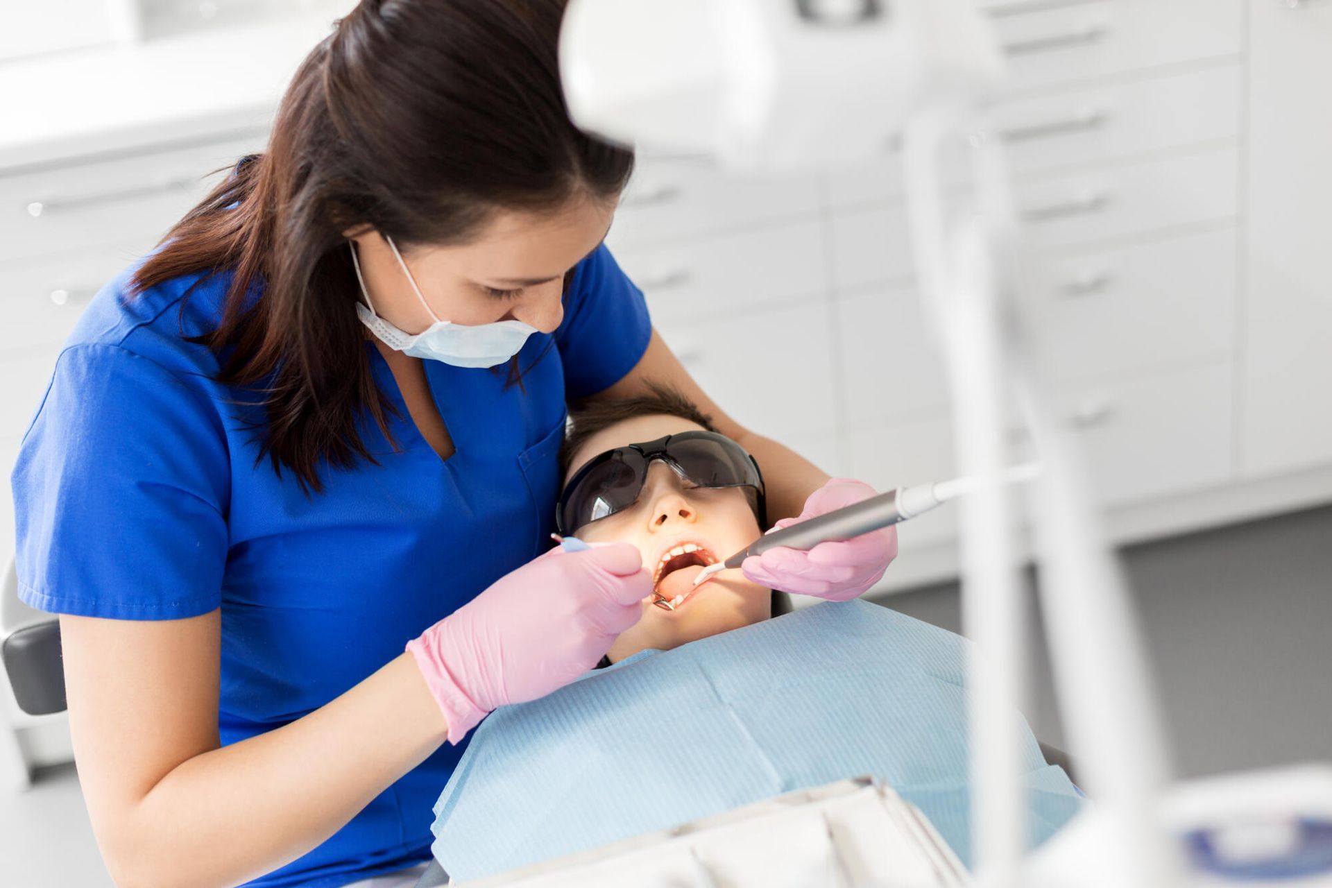 Dentist in blue scrubs treating a patient in a dental chair with a mouth mirror and suction tool