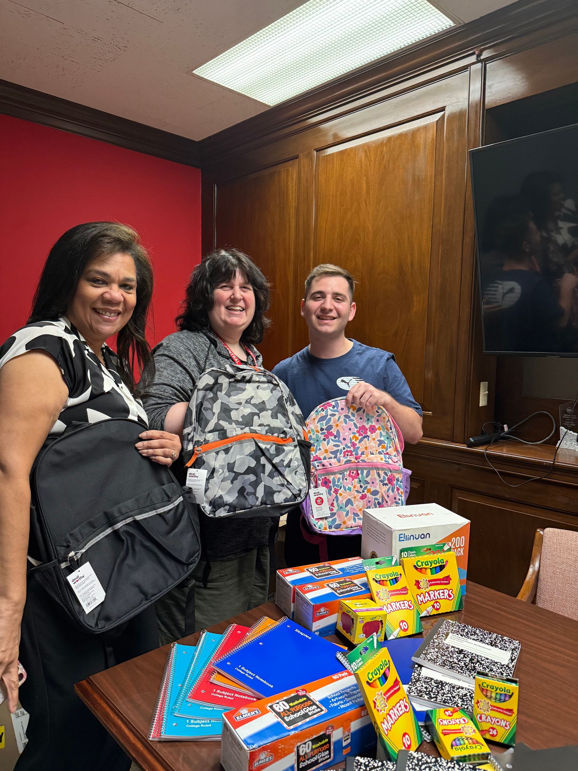 Three people hold backpacks, surrounded by school supplies on a table. Inside a room with red and wood paneling.