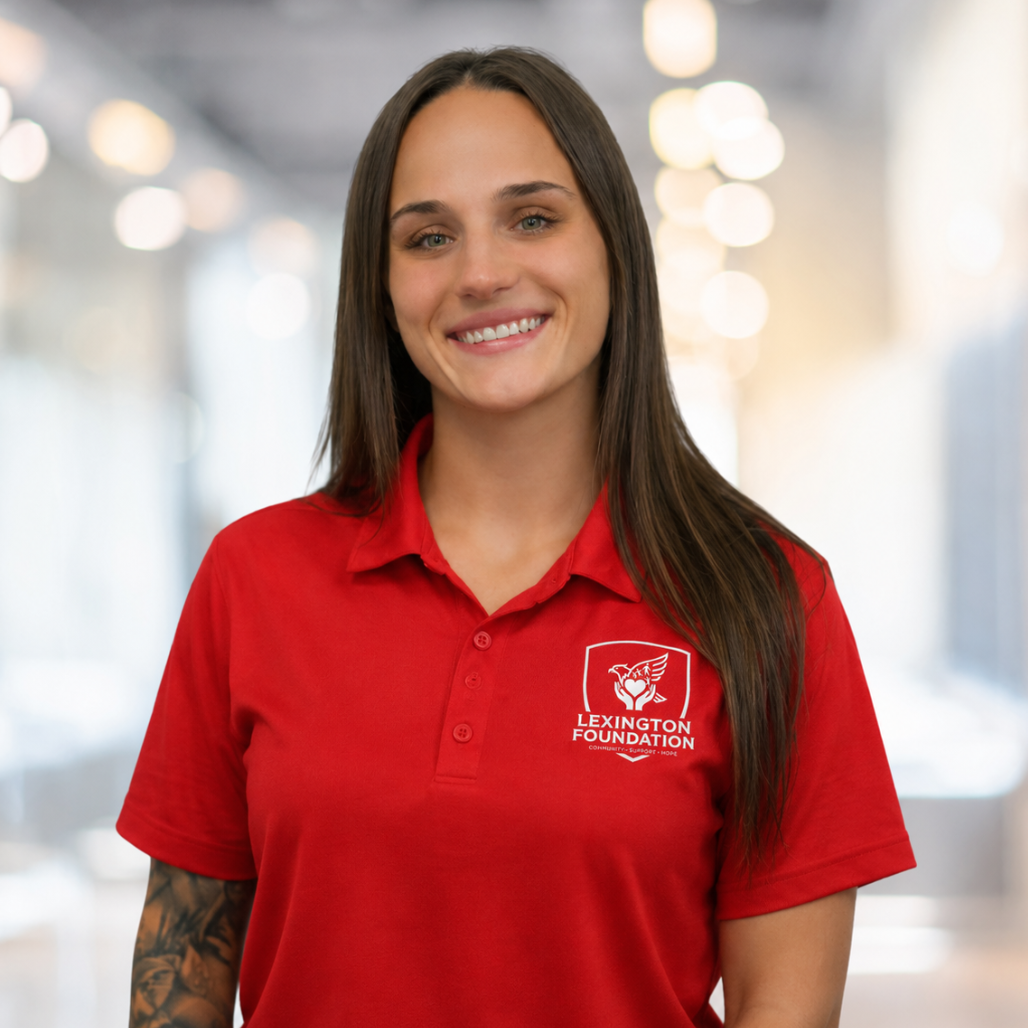 Woman in red polo shirt smiles at the camera, wearing a Lexington logo. Blurred indoor background.