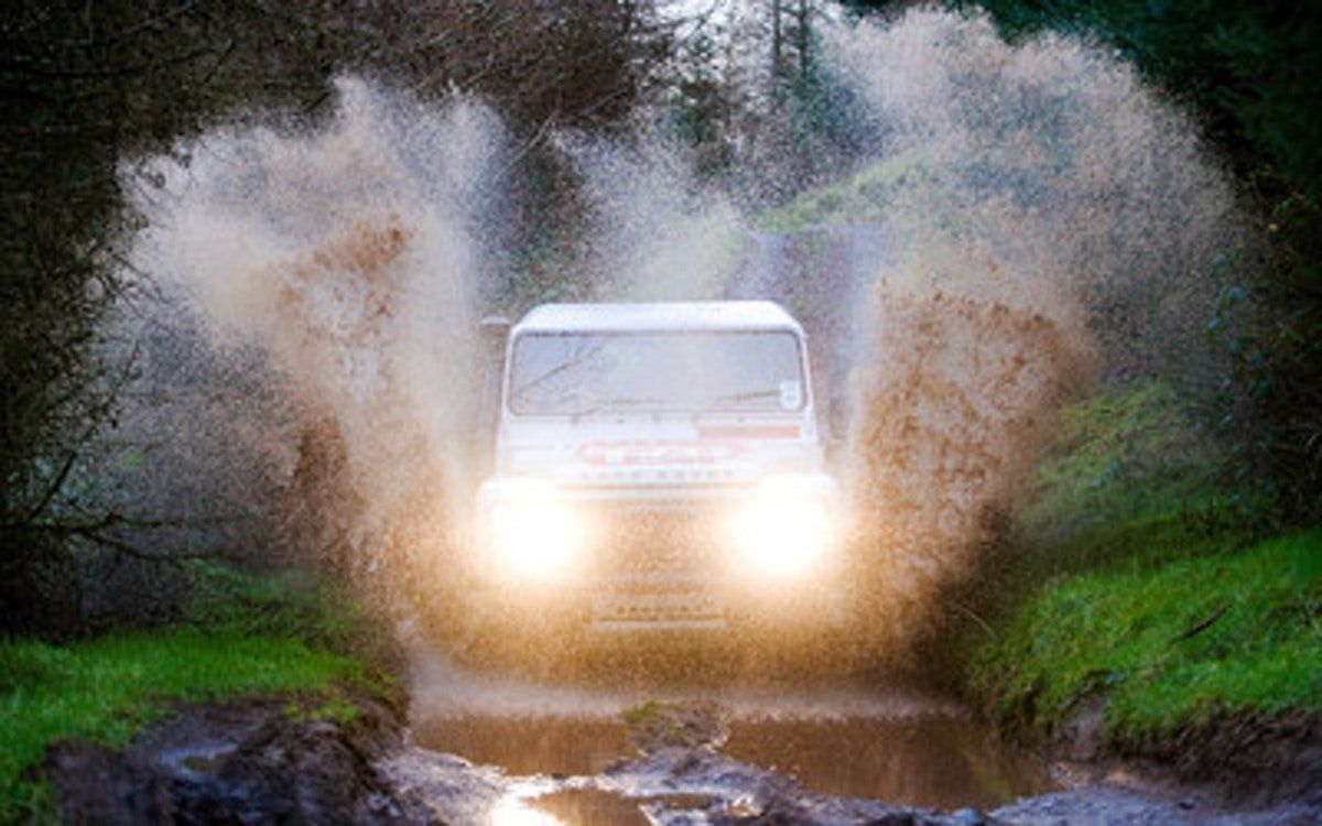 White van splashes through a muddy water crossing on a forest road, headlights on.