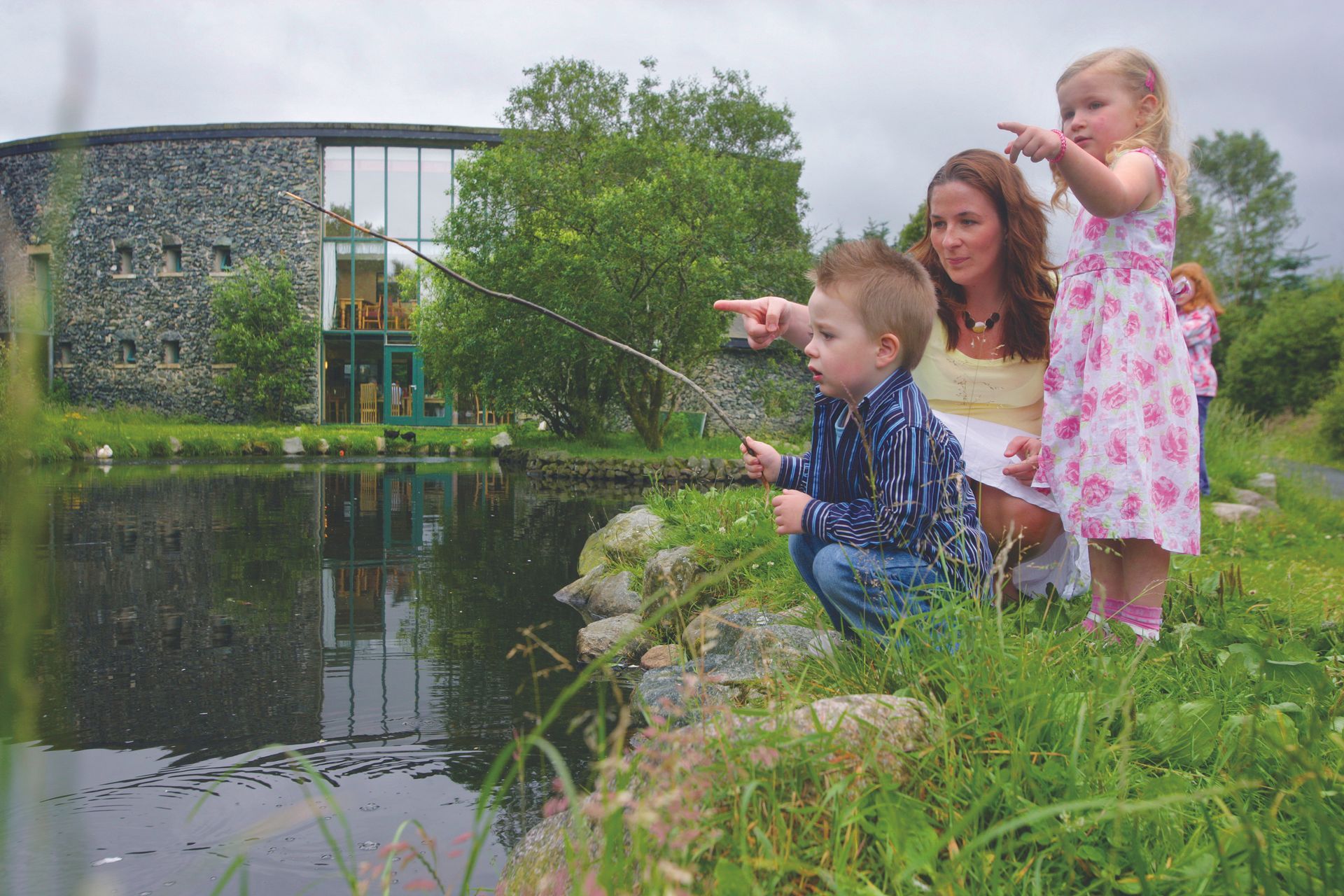Three children sit by a pond, pointing at the water beside a stone building and trees.