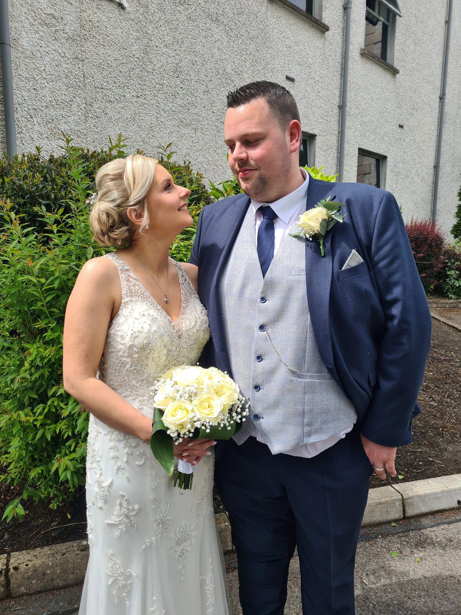 Bride and groom standing outdoors, smiling at each other, holding a bouquet, in wedding attire