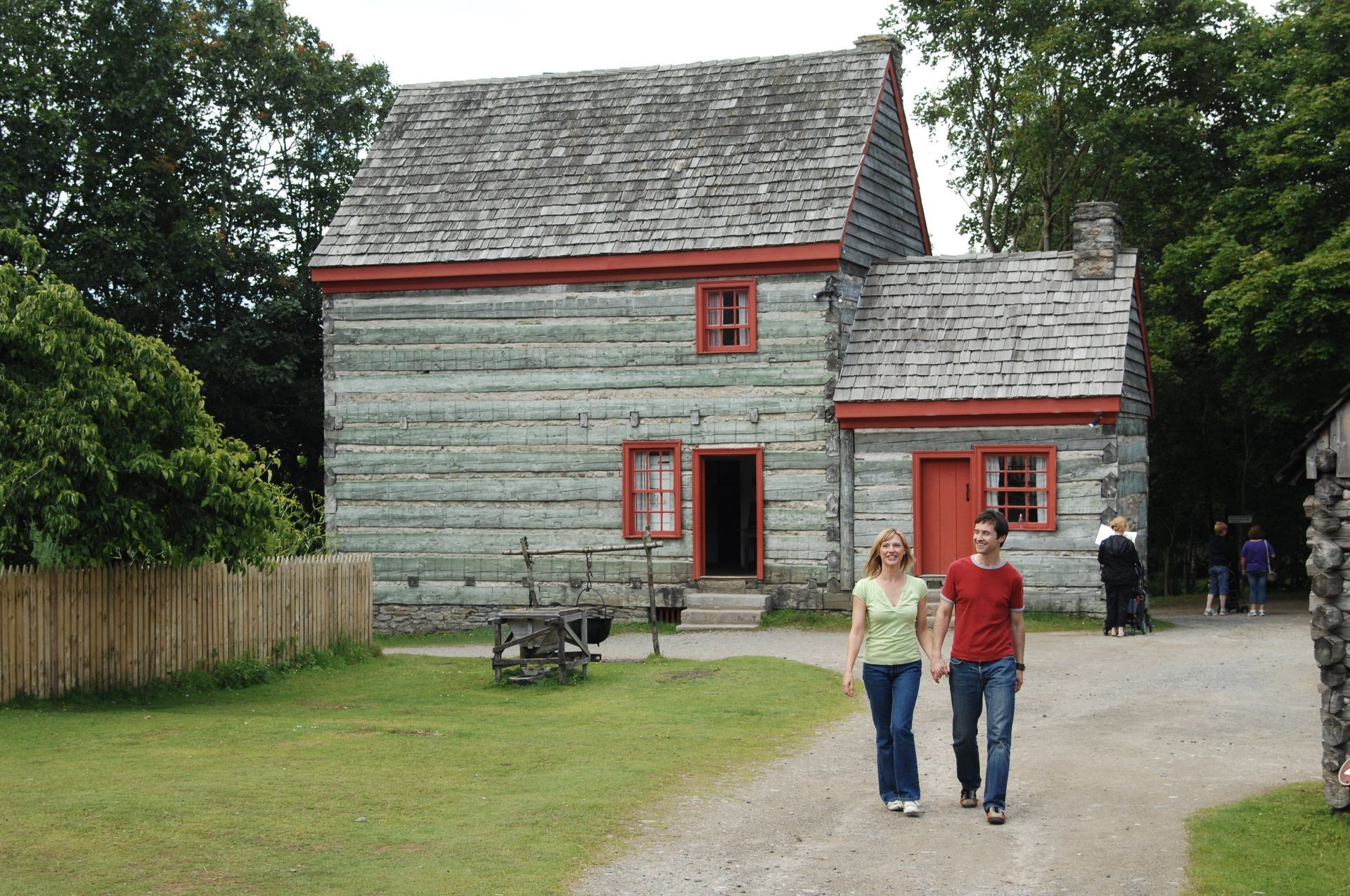 Historic gray wooden house with red trim, two people walking on a gravel path in front.
