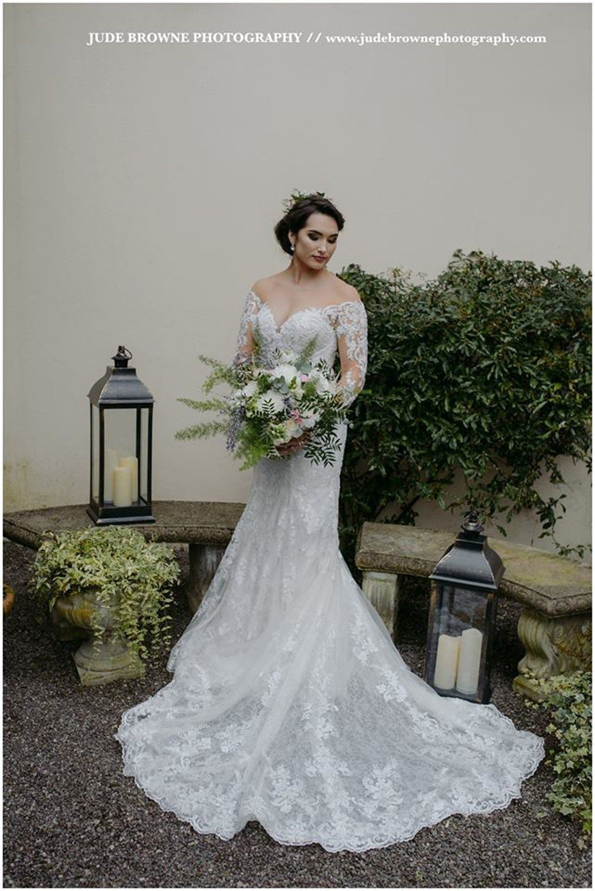 Bride in a white lace gown holding a bouquet beside lanterns and greenery outdoors