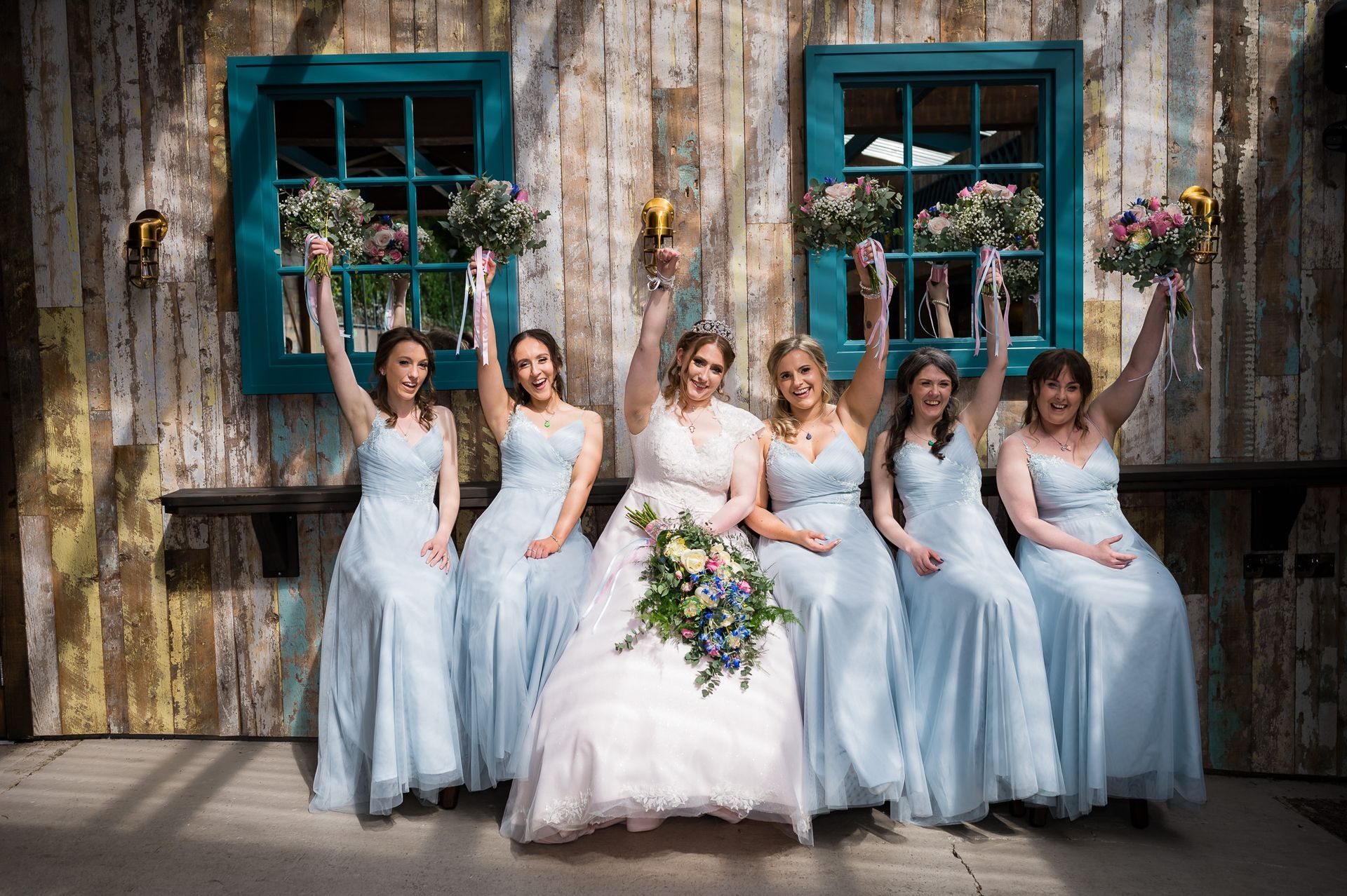Bride and five bridesmaids in light blue dresses pose against a rustic wall with teal windows.