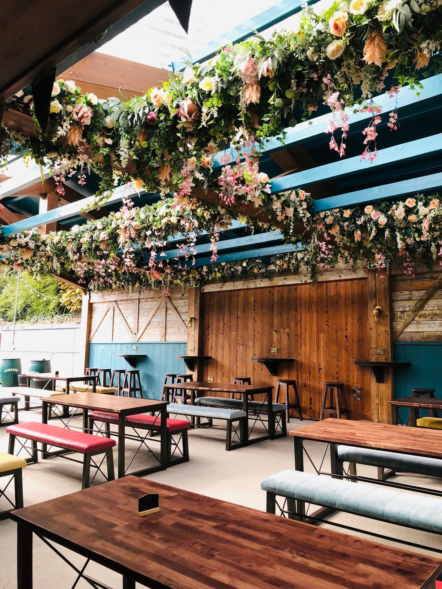 Covered outdoor restaurant patio with wooden tables, benches, and hanging flowers under a blue canopy