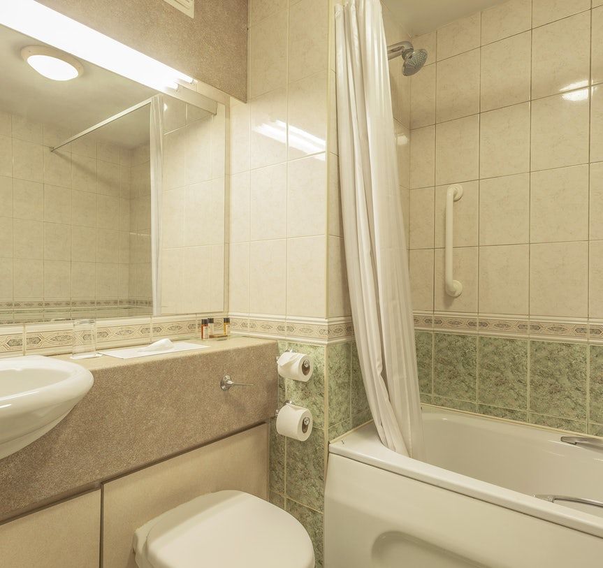 Small beige bathroom with a bathtub, sink, toilet, mirror, and white shower curtain.