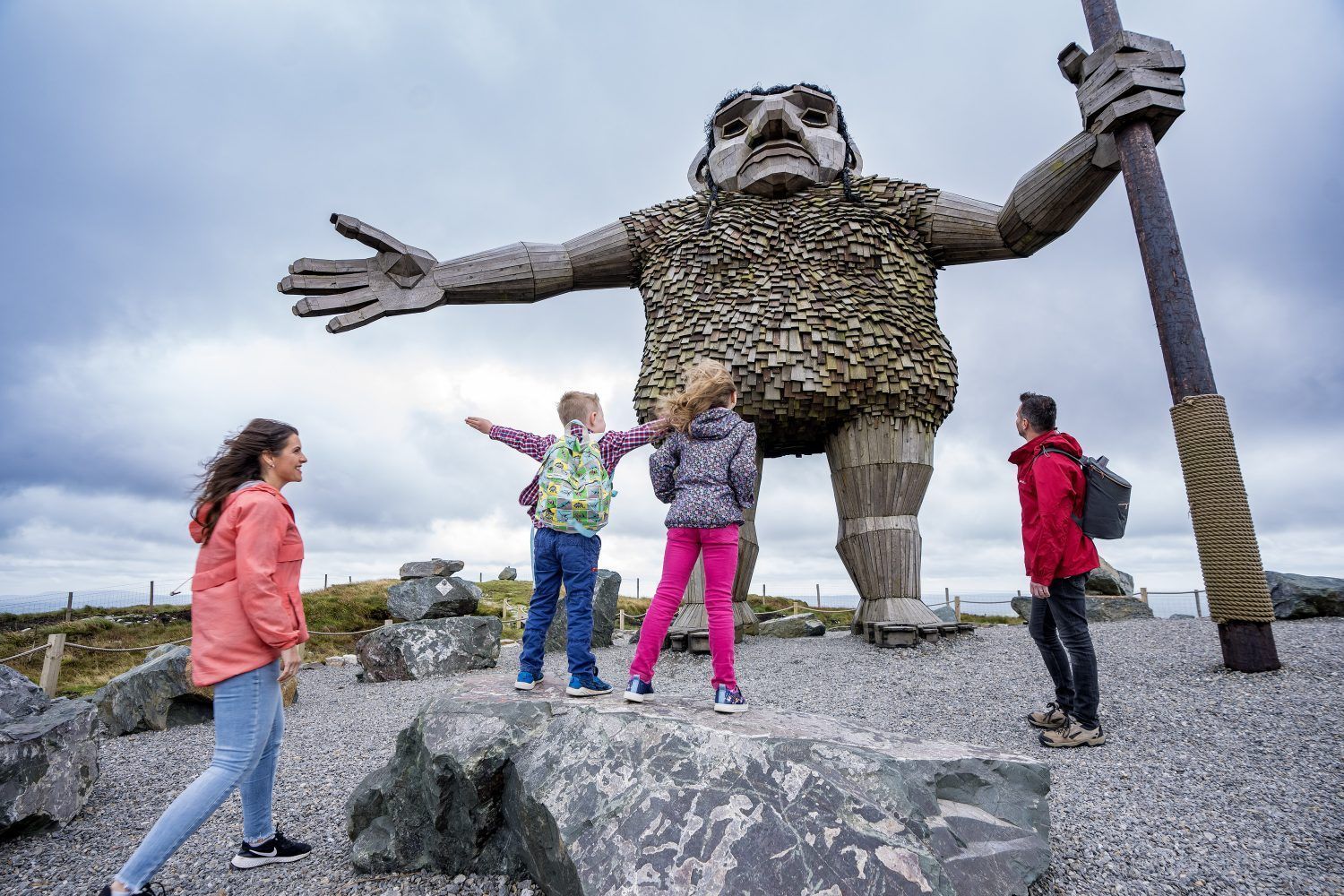 Children pose beside a giant outdoor statue with outstretched arms on rocky ground.