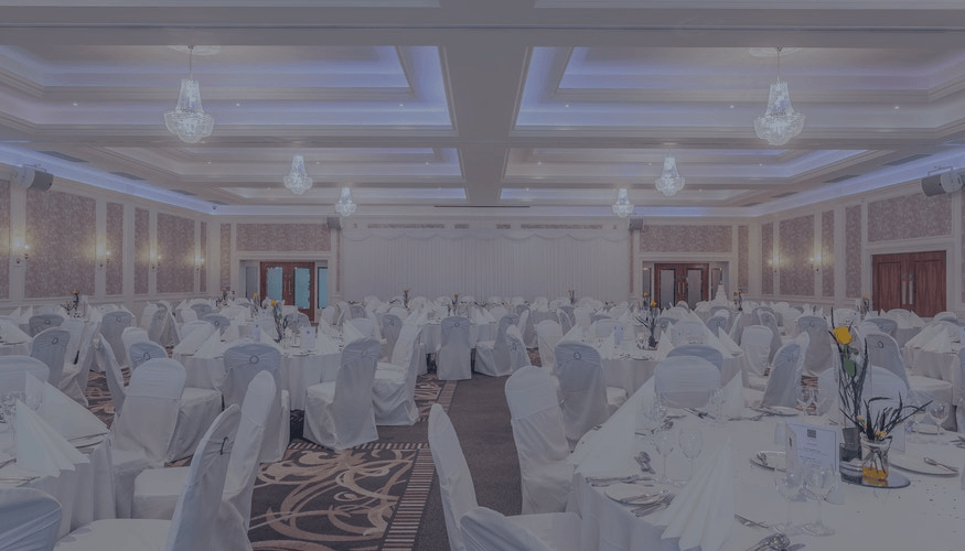 Elegant banquet hall set with round tables, white chair covers, chandeliers, and a stage at the front.