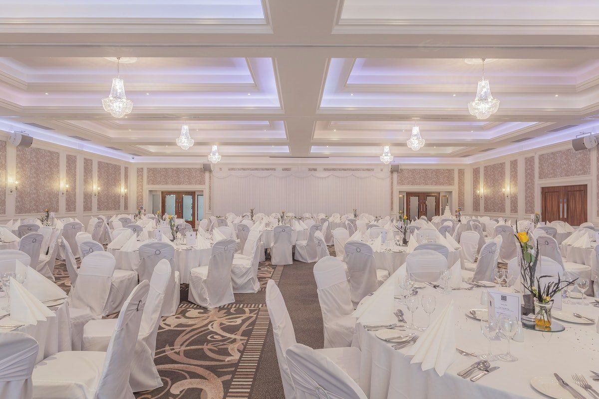 Elegant banquet hall set for a formal event with white-covered chairs and round tables under chandeliers.