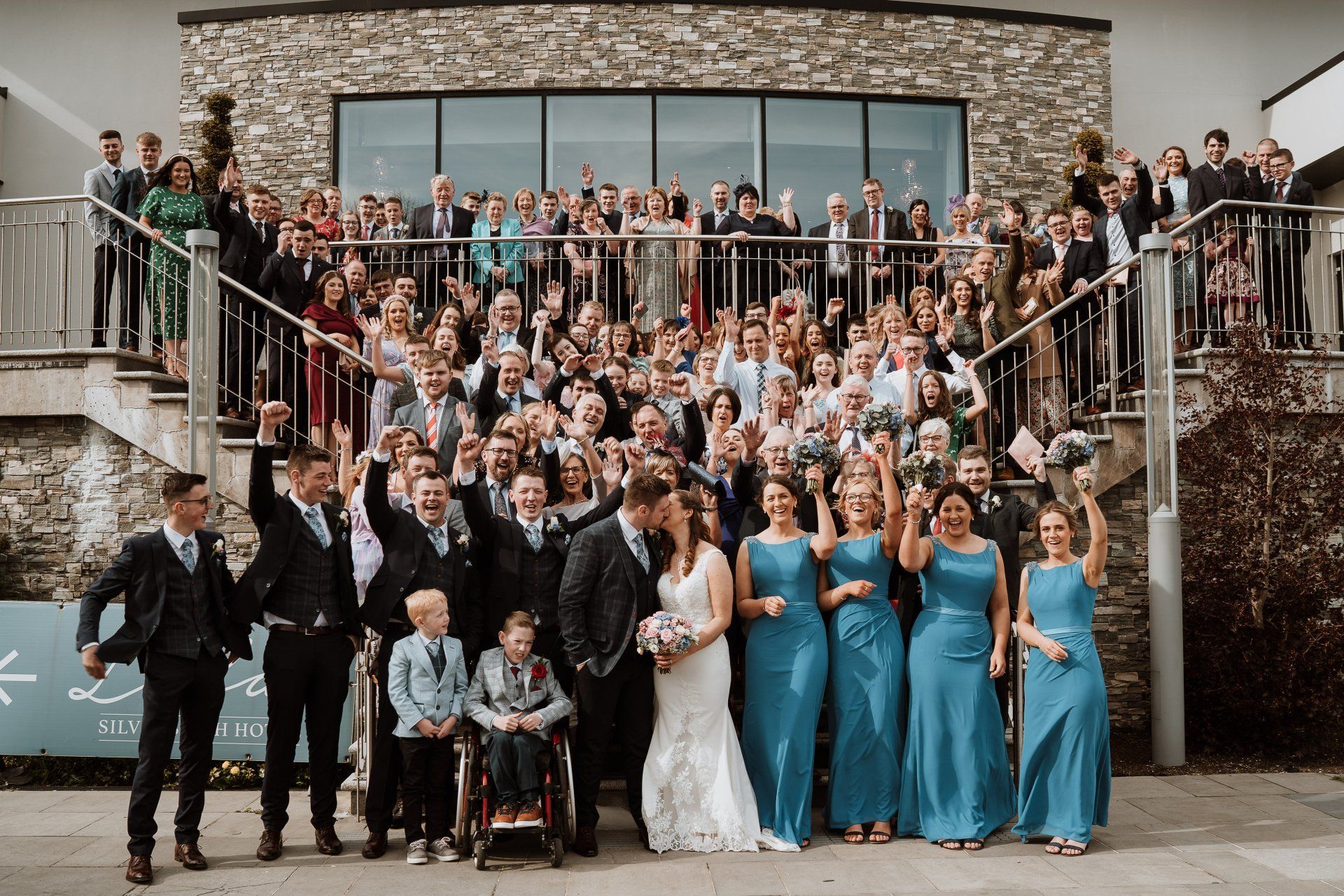 Large wedding party posing on outdoor steps in front of a stone building