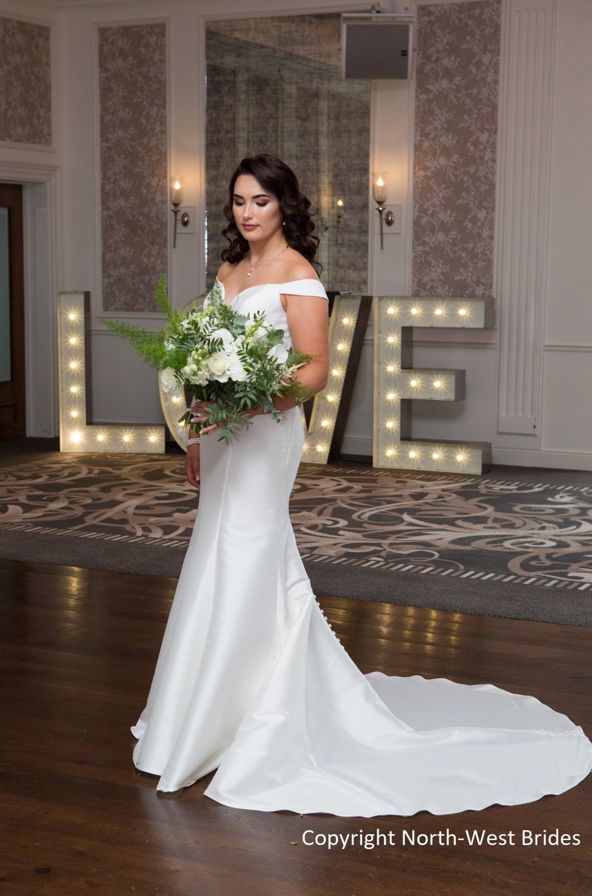 Bride in a white gown holding a bouquet beside illuminated “LOVE” letters indoors